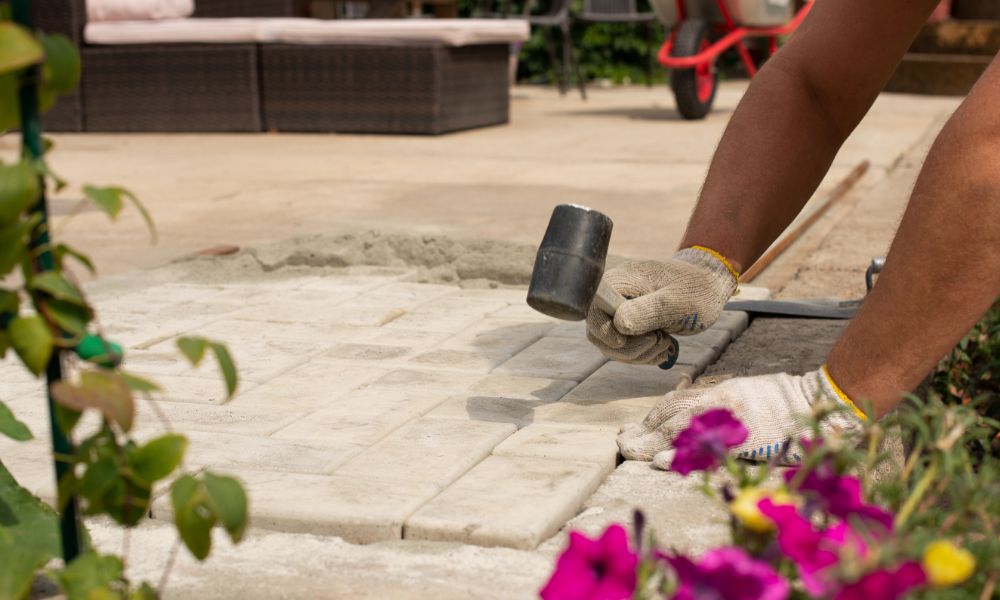 Close-up of a stone mason installing patio pavers with a rubber mallet, surrounded by flowers, in a backyard setting.