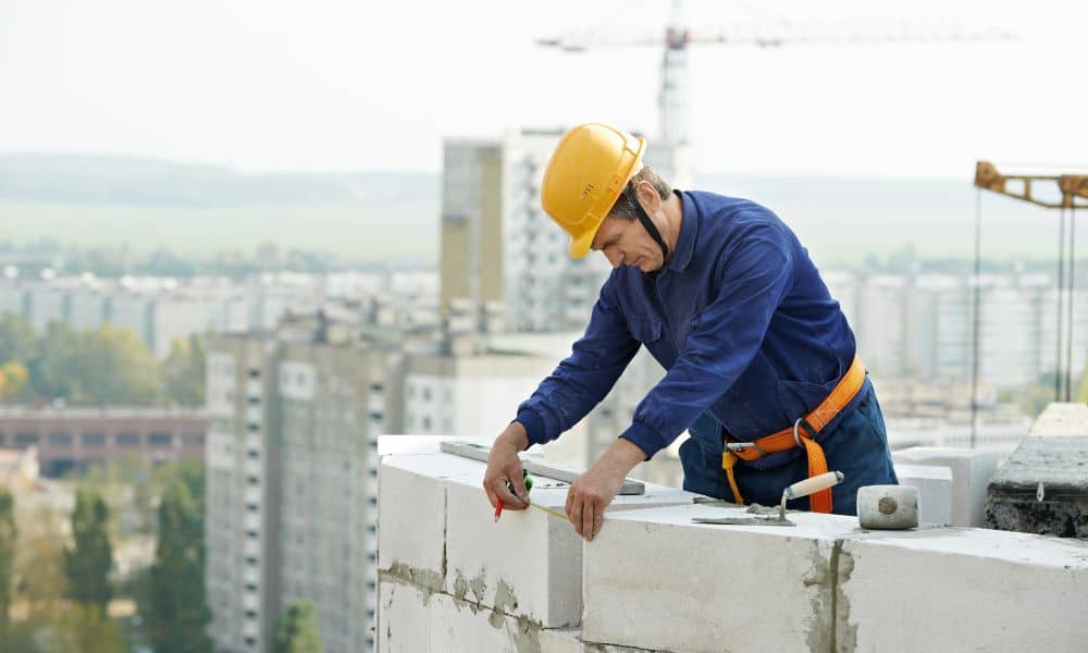 Brick mason using a level to check wall alignment on a high-rise construction site under clear daylight