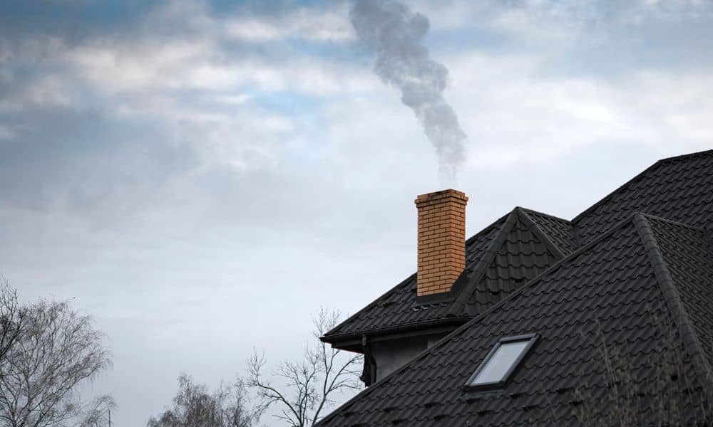 Brick chimney with light smoke rising from a home roof under cloudy skies